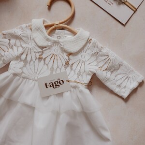 K&ouml;nnte beinhalten: Wei&szlig;es Babykleid mit floraler Spitzenborte und einem Stufenrock, an einem Holzkleiderb&uuml;gel h&auml;ngend. Ein Etikett mit dem Wort "tago" ist angebracht. Ein passendes Stirnband mit floralen Akzenten und dem Text "FROM A LONG TIME AGO..." ist ebenfalls sichtbar.