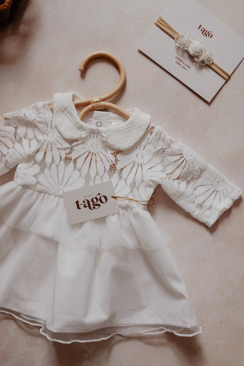 K&ouml;nnte beinhalten: Wei&szlig;es Babykleid mit floraler Spitzenborte und einem Stufenrock, an einem Holzkleiderb&uuml;gel h&auml;ngend. Ein Etikett mit dem Wort "tago" ist angebracht. Ein passendes Stirnband mit floralen Akzenten und dem Text "FROM A LONG TIME AGO..." ist ebenfalls sichtbar.