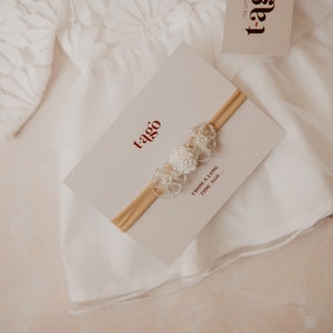 K&ouml;nnte beinhalten: Ein wei&szlig;es Baby-Stirnband mit floralen Verzierungen wird auf einer cremefarbenen Karte pr&auml;sentiert. Die Karte tr&auml;gt den Markennamen "t-ago" in Rot. Das Stirnband befindet sich auf einem cremefarbenen Gummiband. Ein wei&szlig;es Spitzengewand ist im Hintergrund.