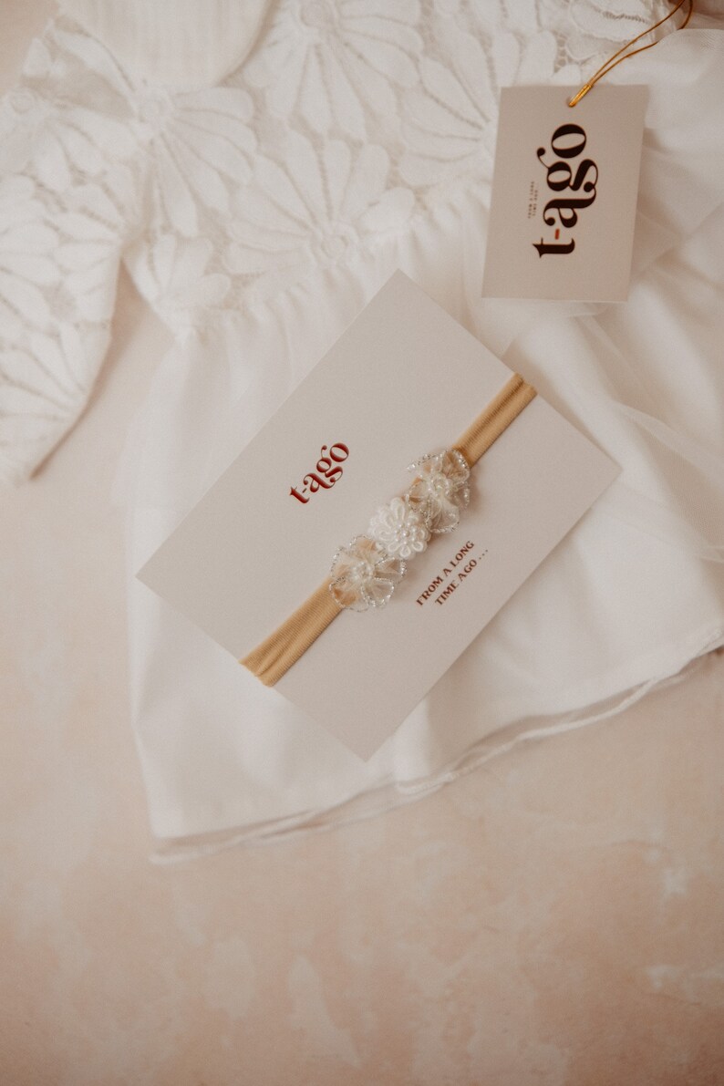 K&ouml;nnte beinhalten: Ein wei&szlig;es Baby-Stirnband mit floralen Verzierungen wird auf einer cremefarbenen Karte pr&auml;sentiert. Die Karte tr&auml;gt den Markennamen "t-ago" in Rot. Das Stirnband befindet sich auf einem cremefarbenen Gummiband. Ein wei&szlig;es Spitzengewand ist im Hintergrund.