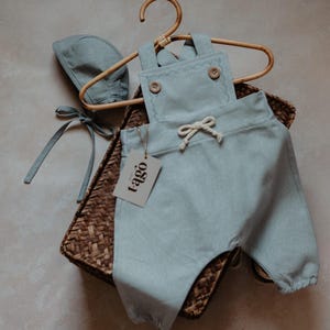 Peut inclure: Ensemble b&eacute;b&eacute; bleu clair compos&eacute; d'une barboteuse et d'un bonnet, avec des boutons en bois et une taille &agrave; cordon. L'ensemble est pr&eacute;sent&eacute; sur un cintre en bois dans un panier en osier. Une &eacute;tiquette avec le mot "tago" est attach&eacute;e.