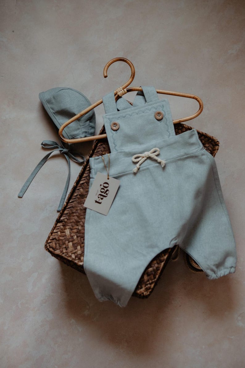Peut inclure: Ensemble b&eacute;b&eacute; bleu clair compos&eacute; d'une barboteuse et d'un bonnet, avec des boutons en bois et une taille &agrave; cordon. L'ensemble est pr&eacute;sent&eacute; sur un cintre en bois dans un panier en osier. Une &eacute;tiquette avec le mot "tago" est attach&eacute;e.