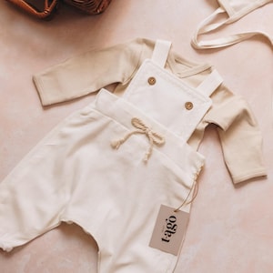 Puede incluir: Un conjunto de ropa de bebé color crema, que incluye un peto con botones de madera y un cordón, un body de manga larga y un gorro. El conjunto tiene una etiqueta que dice "t-ago". Una cesta de mimbre está en el fondo.