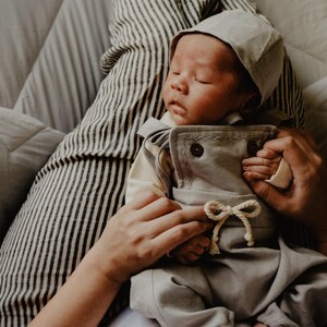 Op de afbeelding: Een slapende baby in een lichtgrijze outfit en bijpassende muts. De outfit heeft een top met knopen en een broek met een trekkoord. De baby wordt vastgehouden door iemand die een gestreept shirt draagt.