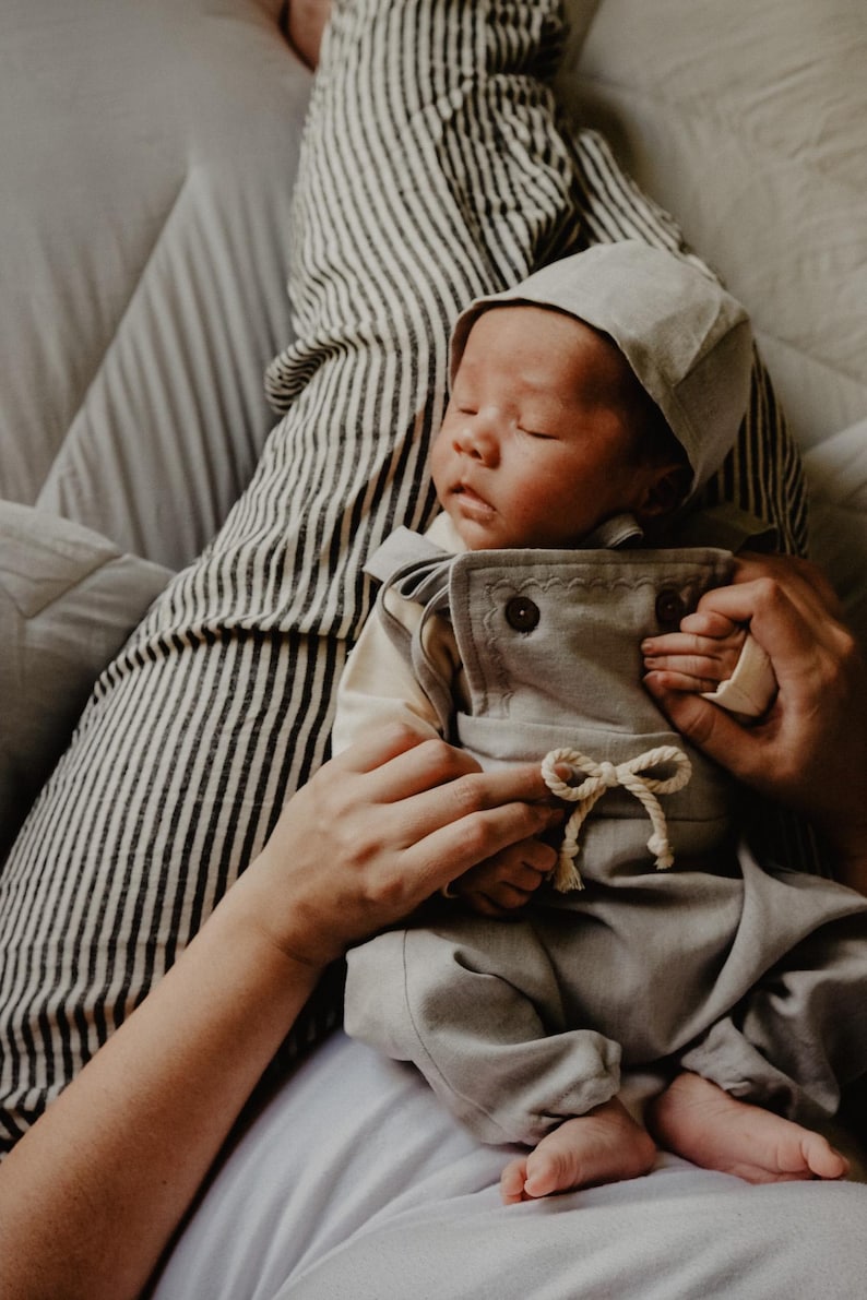Op de afbeelding: Een slapende baby in een lichtgrijze outfit en bijpassende muts. De outfit heeft een top met knopen en een broek met een trekkoord. De baby wordt vastgehouden door iemand die een gestreept shirt draagt.