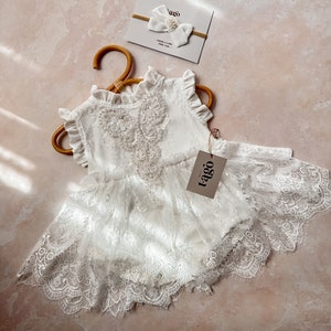 May include: A white baby romper with lace detailing and a matching headband with a bow and pearl accent. The romper is hanging on a wooden hanger. The tag reads "tago".