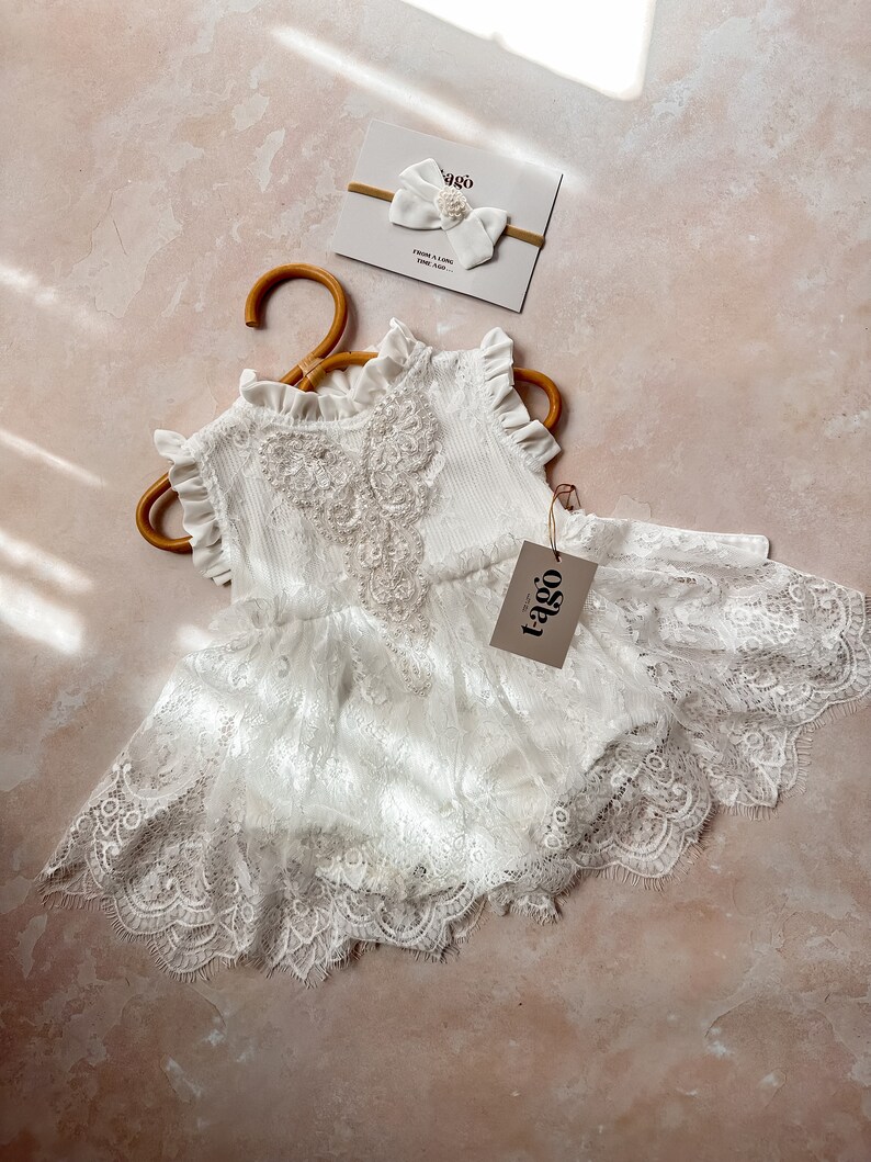 May include: A white baby romper with lace detailing and a matching headband with a bow and pearl accent. The romper is hanging on a wooden hanger. The tag reads "tago".