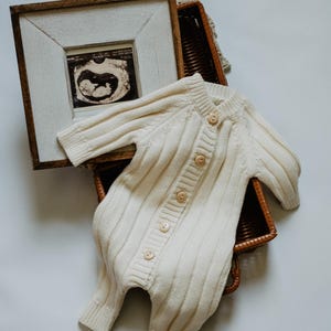May include: Cream-colored knitted baby romper with wooden buttons, resting in a woven basket. A framed ultrasound image is visible, suggesting a baby-themed product. The romper has a ribbed texture and long sleeves.