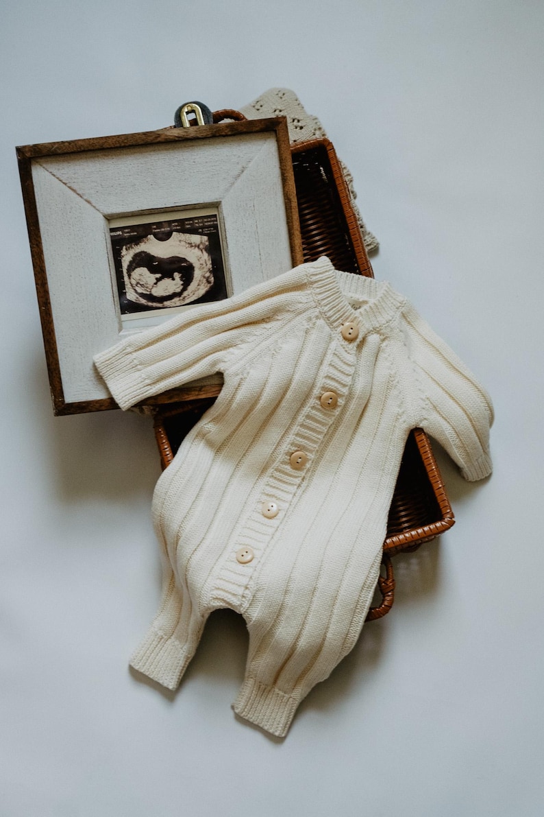 May include: Cream-colored knitted baby romper with wooden buttons, resting in a woven basket. A framed ultrasound image is visible, suggesting a baby-themed product. The romper has a ribbed texture and long sleeves.
