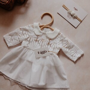 K&ouml;nnte beinhalten: Wei&szlig;es Babykleid mit floraler Spitze am Oberteil und einem mehrlagigen Rock, an einem Holzb&uuml;gel aufgeh&auml;ngt. Ein passendes Stirnband und ein Etikett mit dem Markennamen "tago" sind ebenfalls zu sehen. Das Kleid ist aus weichem, zartem Stoff.