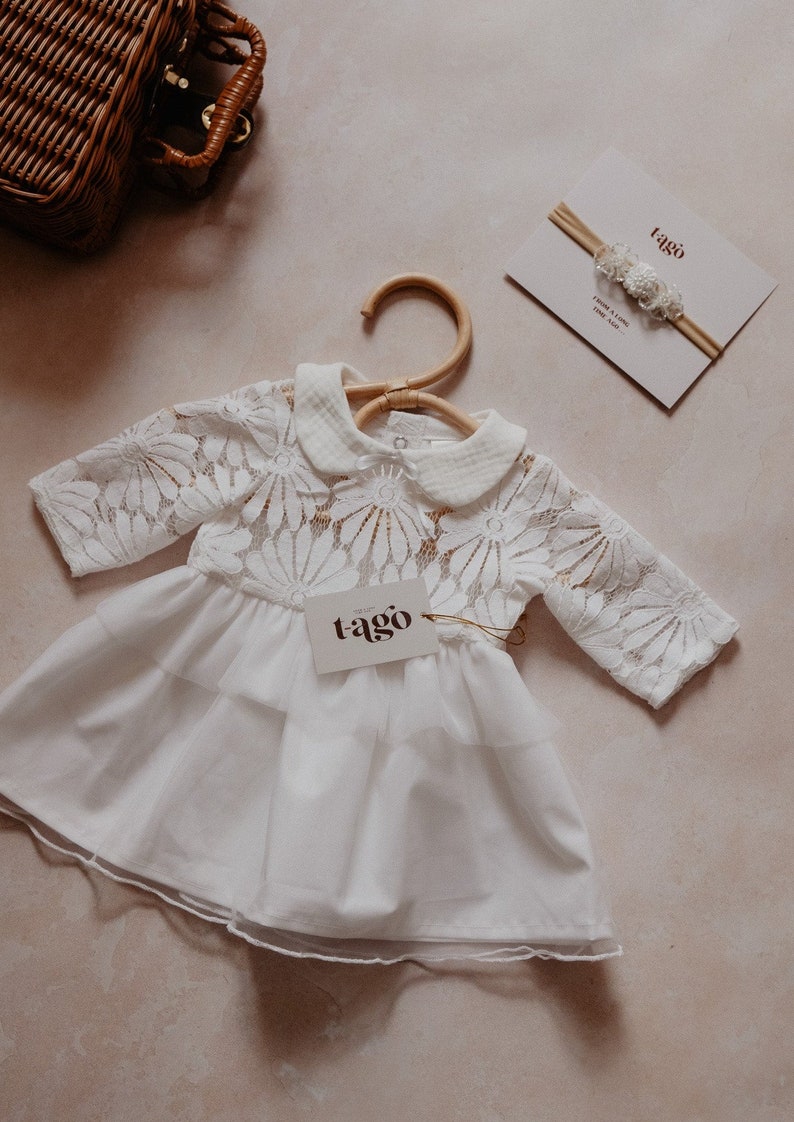K&ouml;nnte beinhalten: Wei&szlig;es Babykleid mit floraler Spitze am Oberteil und einem mehrlagigen Rock, an einem Holzb&uuml;gel aufgeh&auml;ngt. Ein passendes Stirnband und ein Etikett mit dem Markennamen "tago" sind ebenfalls zu sehen. Das Kleid ist aus weichem, zartem Stoff.