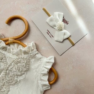 May include: A white baby headband with a large bow and a small floral embellishment, resting on a card with the text "FROM A LONG TIME AGO...". A white lace garment is partially visible, hanging on a wooden hanger.