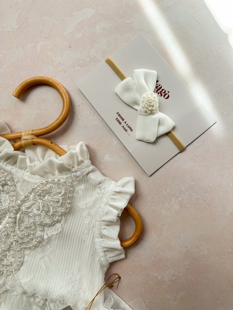 May include: A white baby headband with a large bow and a small floral embellishment, resting on a card with the text "FROM A LONG TIME AGO...". A white lace garment is partially visible, hanging on a wooden hanger.