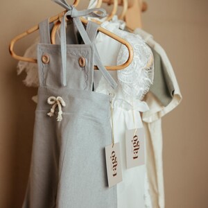 Peut inclure: V&ecirc;tements pour b&eacute;b&eacute;s suspendus &agrave; un portant en bois. Une salopette en lin gris clair avec des boutons en bois et un n&oelig;ud blanc est au premier plan. Une robe blanche avec une bordure en dentelle et une chemise vert clair sont &eacute;galement visibles. Chaque article porte une &eacute;tiquette "tago".