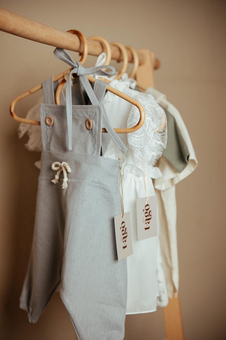 Peut inclure: V&ecirc;tements pour b&eacute;b&eacute;s suspendus &agrave; un portant en bois. Une salopette en lin gris clair avec des boutons en bois et un n&oelig;ud blanc est au premier plan. Une robe blanche avec une bordure en dentelle et une chemise vert clair sont &eacute;galement visibles. Chaque article porte une &eacute;tiquette "tago".