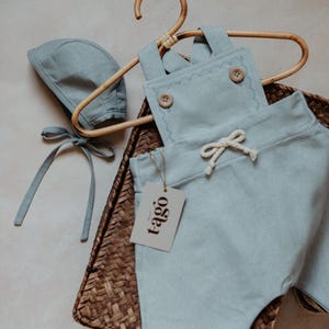 Peut inclure: Ensemble de v&ecirc;tements pour b&eacute;b&eacute; bleu clair, comprenant une salopette et un bonnet, pr&eacute;sent&eacute; sur un cintre en bois. La salopette a des bords festonn&eacute;s d&eacute;coratifs et des boutons en bois. Une &eacute;tiquette avec le mot "tago" est attach&eacute;e. L'ensemble est dans un panier tress&eacute;.