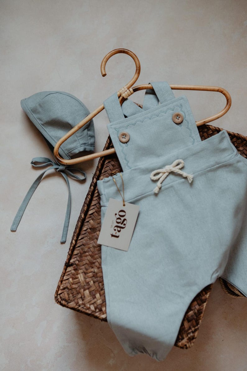Peut inclure: Ensemble de v&ecirc;tements pour b&eacute;b&eacute; bleu clair, comprenant une salopette et un bonnet, pr&eacute;sent&eacute; sur un cintre en bois. La salopette a des bords festonn&eacute;s d&eacute;coratifs et des boutons en bois. Une &eacute;tiquette avec le mot "tago" est attach&eacute;e. L'ensemble est dans un panier tress&eacute;.