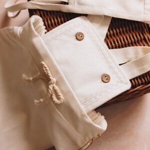 May include: Cream-colored baby outfit set with a bonnet and overalls, featuring wooden buttons and a rope tie. The set is placed on a woven basket, with a tag that reads "tago".