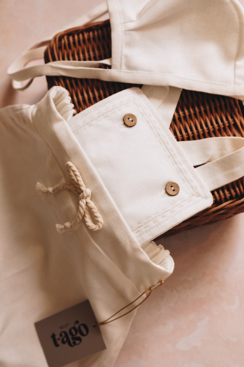 May include: Cream-colored baby outfit set with a bonnet and overalls, featuring wooden buttons and a rope tie. The set is placed on a woven basket, with a tag that reads "tago".