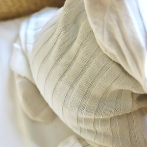 May include: Close-up of a cream-colored, ribbed knit fabric. The fabric is soft and appears to be a blanket or swaddling cloth. The texture is visible, with vertical ribbing creating a subtle pattern. The overall aesthetic is simple and cozy.