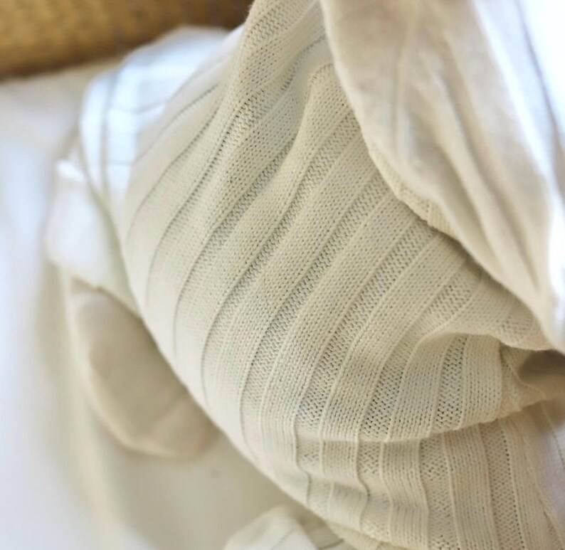 May include: Close-up of a cream-colored, ribbed knit fabric. The fabric is soft and appears to be a blanket or swaddling cloth. The texture is visible, with vertical ribbing creating a subtle pattern. The overall aesthetic is simple and cozy.