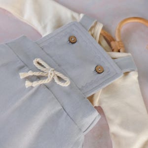 Op de afbeelding: Lichtblauwe babybroek met een trekkoord en een bijpassende top. De broek heeft een cr&egrave;mekleurig trekkoord en een decoratieve flap met twee houten knopen. De top is cr&egrave;mekleurig en heeft grijze afwerking.