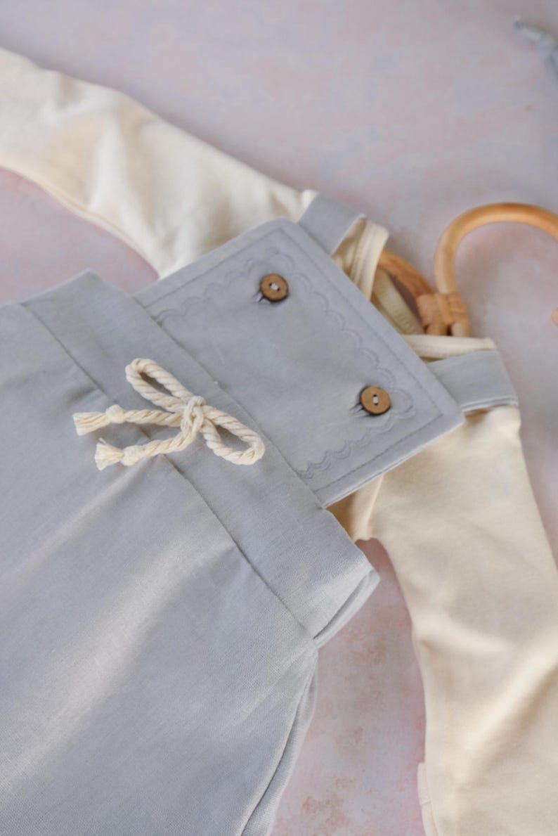 Op de afbeelding: Lichtblauwe babybroek met een trekkoord en een bijpassende top. De broek heeft een cr&egrave;mekleurig trekkoord en een decoratieve flap met twee houten knopen. De top is cr&egrave;mekleurig en heeft grijze afwerking.
