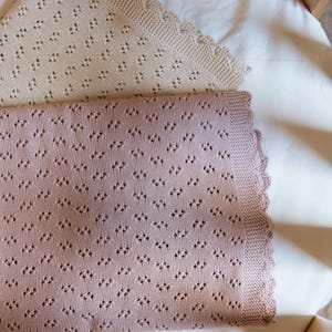 May include: Two knitted baby blankets. One is a light beige color, and the other is a dusty rose color. Both blankets have a diamond-shaped pattern and a scalloped edge. The blankets are lying on a white sheet.