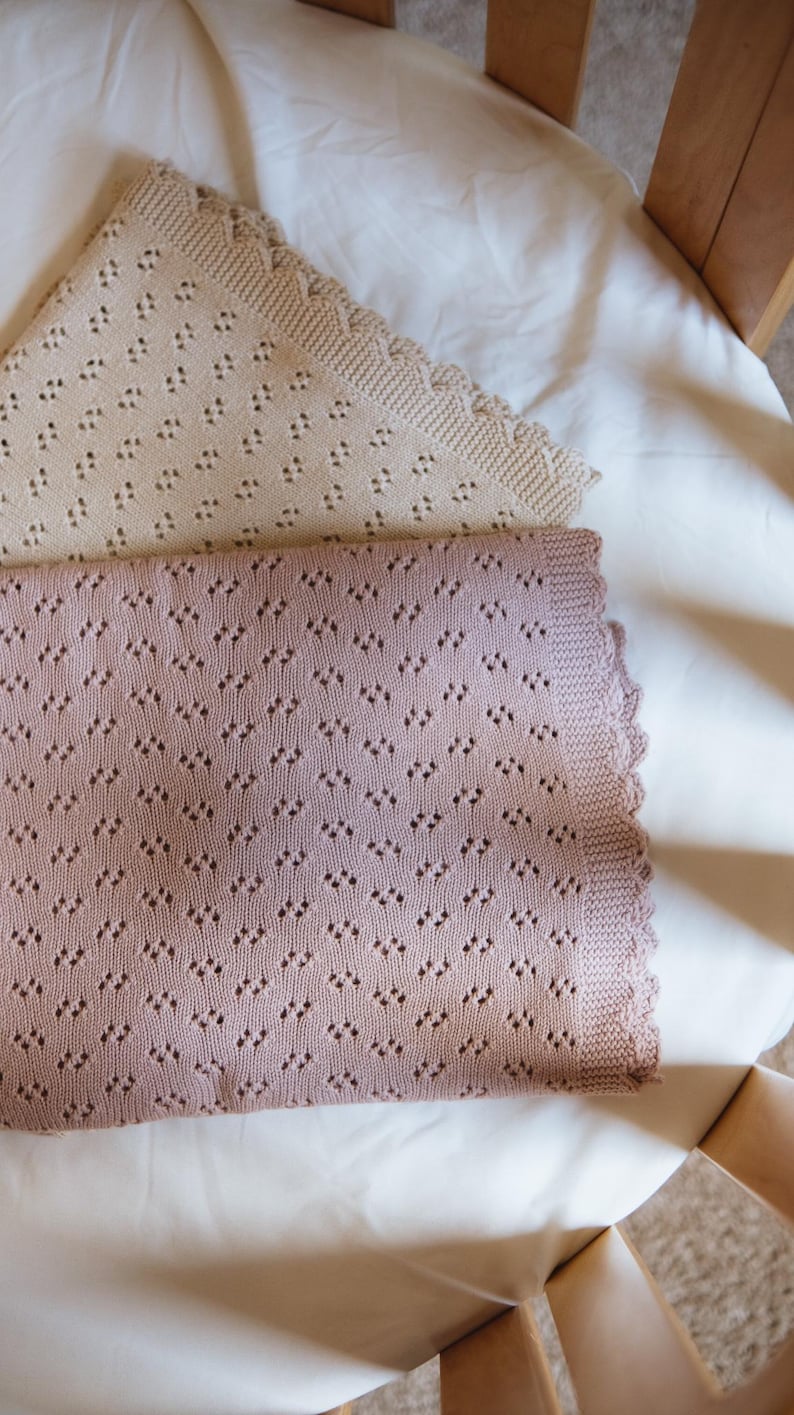 May include: Two knitted baby blankets. One is a light beige color, and the other is a dusty rose color. Both blankets have a diamond-shaped pattern and a scalloped edge. The blankets are lying on a white sheet.
