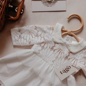K&ouml;nnte beinhalten: Ein wei&szlig;es Babykleid mit floralen Spitzen&auml;rmeln und -oberteil, das an einem Holzkleiderb&uuml;gel h&auml;ngt. Ein passendes Stirnband mit floralen Akzenten und ein Etikett mit dem Markennamen "tago" sind ebenfalls sichtbar. Der Text "FROM A LONG TIME AGO..." steht auf der Karte.