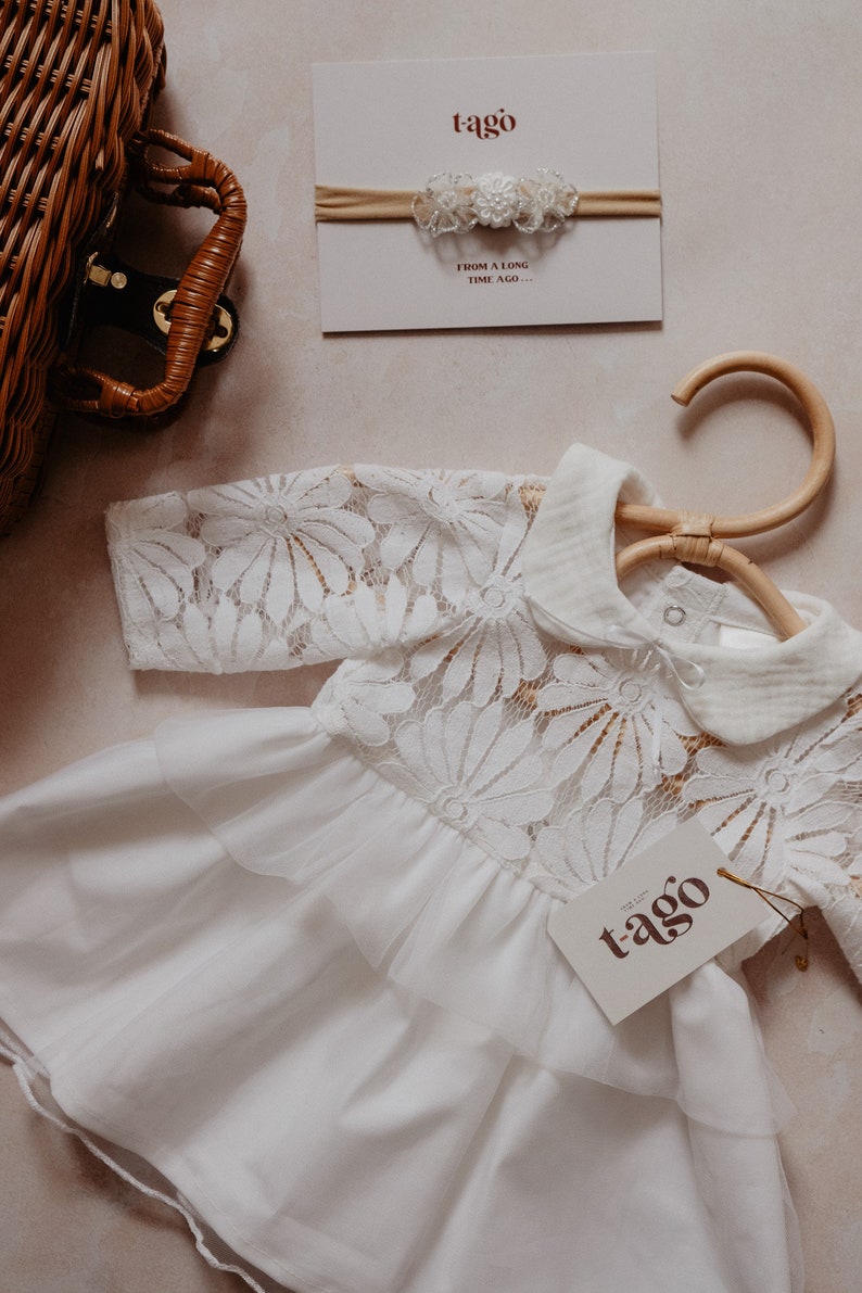 K&ouml;nnte beinhalten: Ein wei&szlig;es Babykleid mit floralen Spitzen&auml;rmeln und -oberteil, das an einem Holzkleiderb&uuml;gel h&auml;ngt. Ein passendes Stirnband mit floralen Akzenten und ein Etikett mit dem Markennamen "tago" sind ebenfalls sichtbar. Der Text "FROM A LONG TIME AGO..." steht auf der Karte.