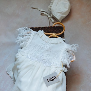 Puede incluir: Un vestido de bebé blanco con detalles de encaje y un gorro a juego descansan en una cesta de mimbre. El vestido tiene mangas con volantes y una etiqueta que dice "tago/". El fondo es de un color suave y neutro.