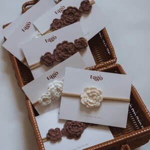 May include: A woven basket holds several cards displaying crochet flower headbands. The headbands are in shades of brown and white, with the word "tago" printed on the cards. The headbands are made of yarn and are attached to a thin, neutral-colored band.