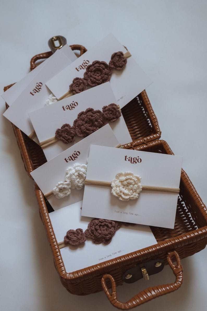 May include: A woven basket holds several cards displaying crochet flower headbands. The headbands are in shades of brown and white, with the word "tago" printed on the cards. The headbands are made of yarn and are attached to a thin, neutral-colored band.
