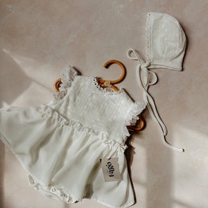 May include: A white baby romper with a ruffled skirt and eyelet bodice, hanging on a wooden hanger. A matching bonnet with a lace trim and tie is next to the romper. The tag reads "t-ago".
