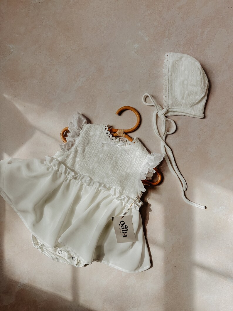 May include: A white baby romper with a ruffled skirt and eyelet bodice, hanging on a wooden hanger. A matching bonnet with a lace trim and tie is next to the romper. The tag reads "t-ago".