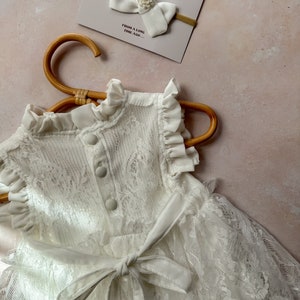 May include: A white lace dress with a matching bow headband. The dress features a high collar, ruffled sleeves, and a tie-waist. The headband has a large bow with a pearl accent. A card with the text "ago" and "FROM A LONG TIME AGO..." is also visible.