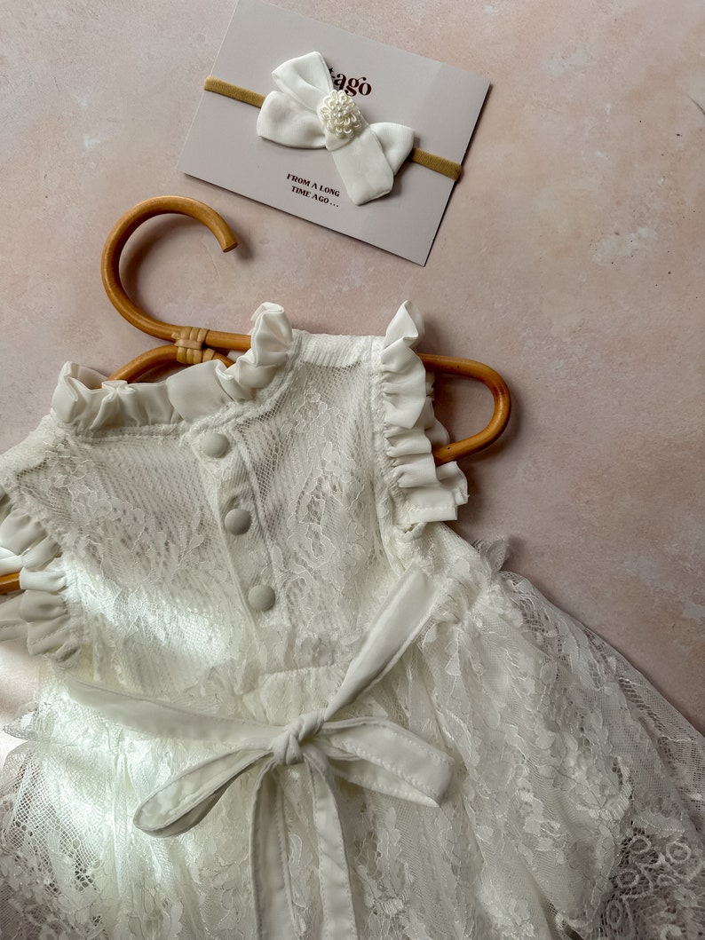 May include: A white lace dress with a matching bow headband. The dress features a high collar, ruffled sleeves, and a tie-waist. The headband has a large bow with a pearl accent. A card with the text "ago" and "FROM A LONG TIME AGO..." is also visible.