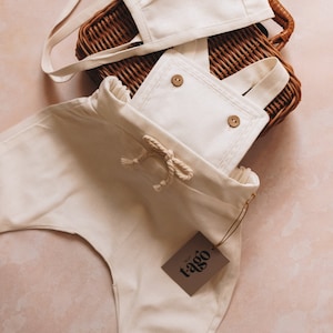 May include: Cream-colored baby outfit set, including overalls with wooden buttons and a matching bonnet. The outfit is made of soft fabric and features a drawstring waist. A brown tag with the text "t-ago" is attached.