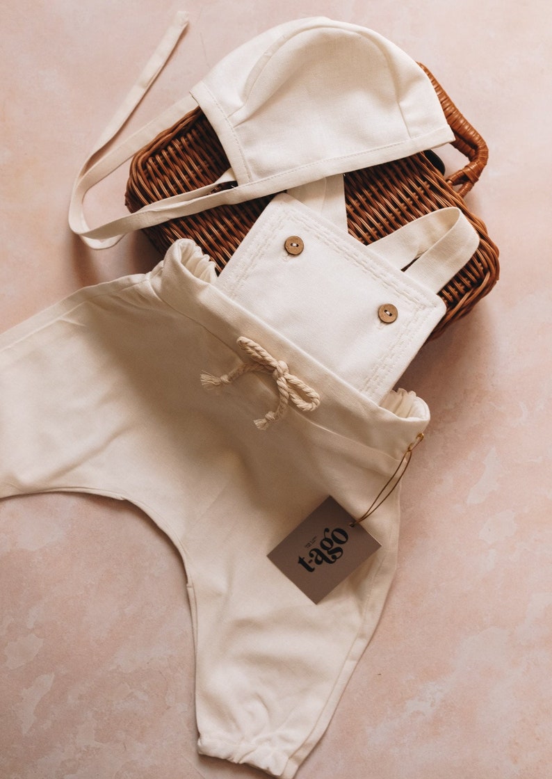 May include: Cream-colored baby outfit set, including overalls with wooden buttons and a matching bonnet. The outfit is made of soft fabric and features a drawstring waist. A brown tag with the text "t-ago" is attached.