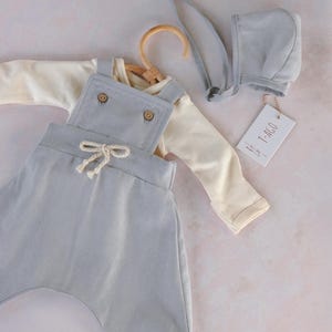 Op de afbeelding: Een babykleding set in zachte pastelkleuren. De set bevat een lichtblauwe overall met houten knopen, een cr&egrave;mekleurig shirt met lange mouwen en een bijpassend mutsje. Een label met de tekst "T-AGO" is bevestigd.
