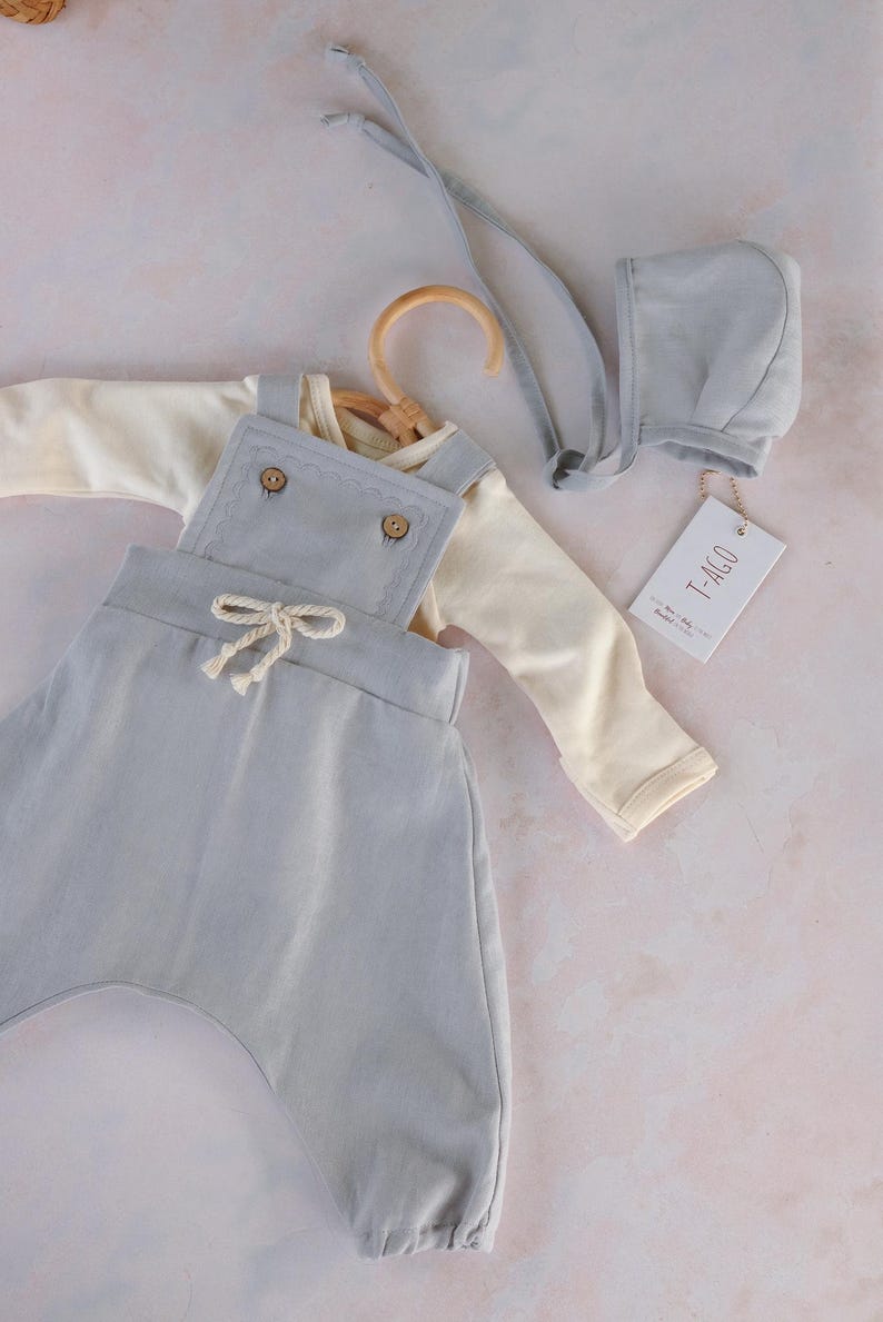 Op de afbeelding: Een babykleding set in zachte pastelkleuren. De set bevat een lichtblauwe overall met houten knopen, een cr&egrave;mekleurig shirt met lange mouwen en een bijpassend mutsje. Een label met de tekst "T-AGO" is bevestigd.