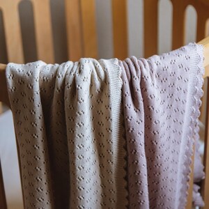 May include: A soft, knitted baby blanket in shades of cream and mauve, draped over a wooden crib. The blanket features a delicate, openwork pattern and a scalloped edge, creating a cozy and inviting aesthetic. The crib is made of light-colored wood.