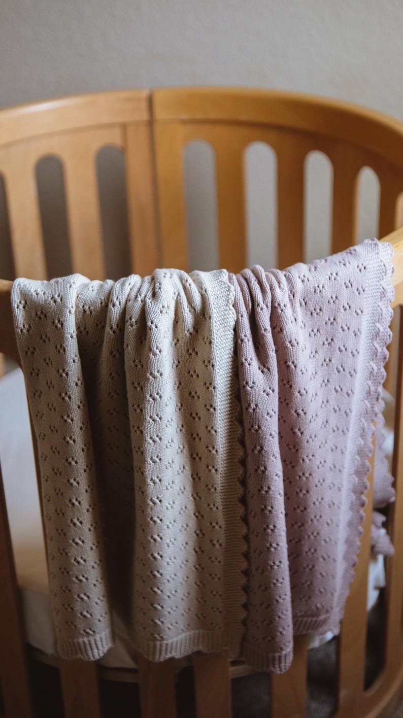 May include: A soft, knitted baby blanket in shades of cream and mauve, draped over a wooden crib. The blanket features a delicate, openwork pattern and a scalloped edge, creating a cozy and inviting aesthetic. The crib is made of light-colored wood.