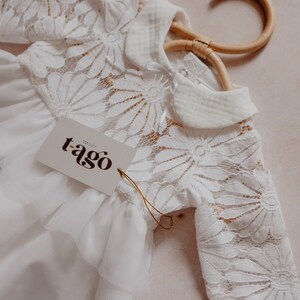 K&ouml;nnte beinhalten: Wei&szlig;er Baby-Strampler mit floralen Spitzen&auml;rmeln und -oberteil, an einem Holzb&uuml;gel h&auml;ngend. Der Strampler hat einen Bubikragen und ein Etikett mit der Aufschrift "t-ago". Das Kleidungsst&uuml;ck hat einen ger&uuml;schten Rock.