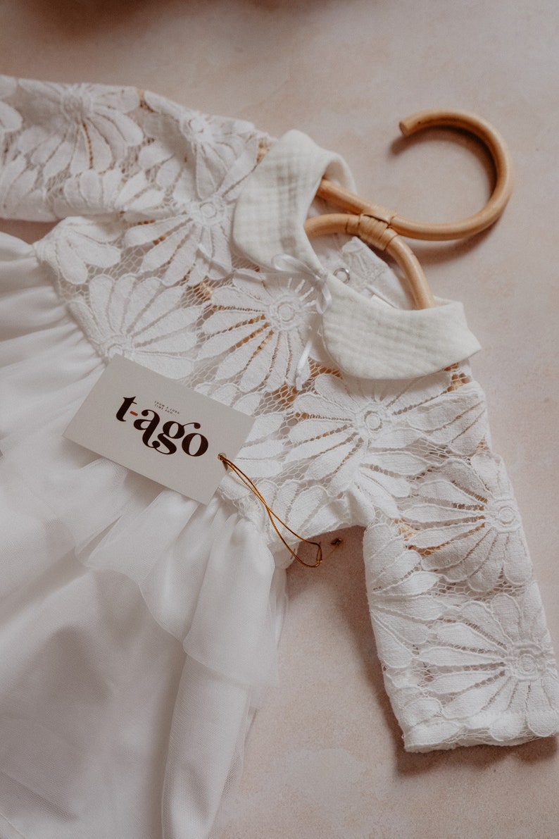 K&ouml;nnte beinhalten: Wei&szlig;er Baby-Strampler mit floralen Spitzen&auml;rmeln und -oberteil, an einem Holzb&uuml;gel h&auml;ngend. Der Strampler hat einen Bubikragen und ein Etikett mit der Aufschrift "t-ago". Das Kleidungsst&uuml;ck hat einen ger&uuml;schten Rock.