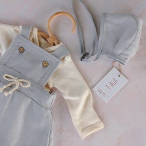 Op de afbeelding: Een baby-outfitset in zachte pastelkleuren. De set bevat een cr&egrave;mekleurig shirt met lange mouwen, een lichtblauwe tuinbroek met houten knopen en een bijpassend mutsje. Een houten kleerhanger en een label met tekst zijn ook inbegrepen.