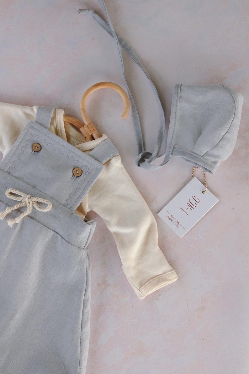 Op de afbeelding: Een baby-outfitset in zachte pastelkleuren. De set bevat een cr&egrave;mekleurig shirt met lange mouwen, een lichtblauwe tuinbroek met houten knopen en een bijpassend mutsje. Een houten kleerhanger en een label met tekst zijn ook inbegrepen.