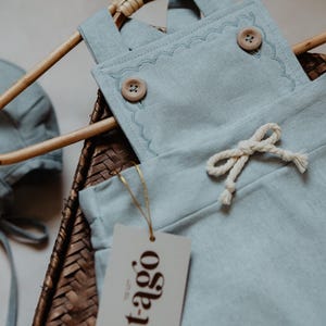 Peut inclure: Un ensemble de v&ecirc;tements pour b&eacute;b&eacute; bleu clair, comprenant une salopette avec des bords festonn&eacute;s et des boutons en bois, et un bonnet assorti. L'ensemble est pr&eacute;sent&eacute; sur un cintre en osier avec une &eacute;tiquette "t-ago".