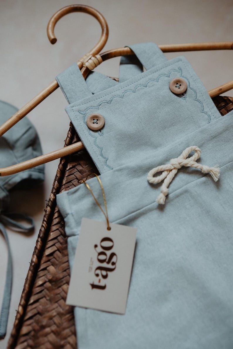 Peut inclure: Un ensemble de v&ecirc;tements pour b&eacute;b&eacute; bleu clair, comprenant une salopette avec des bords festonn&eacute;s et des boutons en bois, et un bonnet assorti. L'ensemble est pr&eacute;sent&eacute; sur un cintre en osier avec une &eacute;tiquette "t-ago".